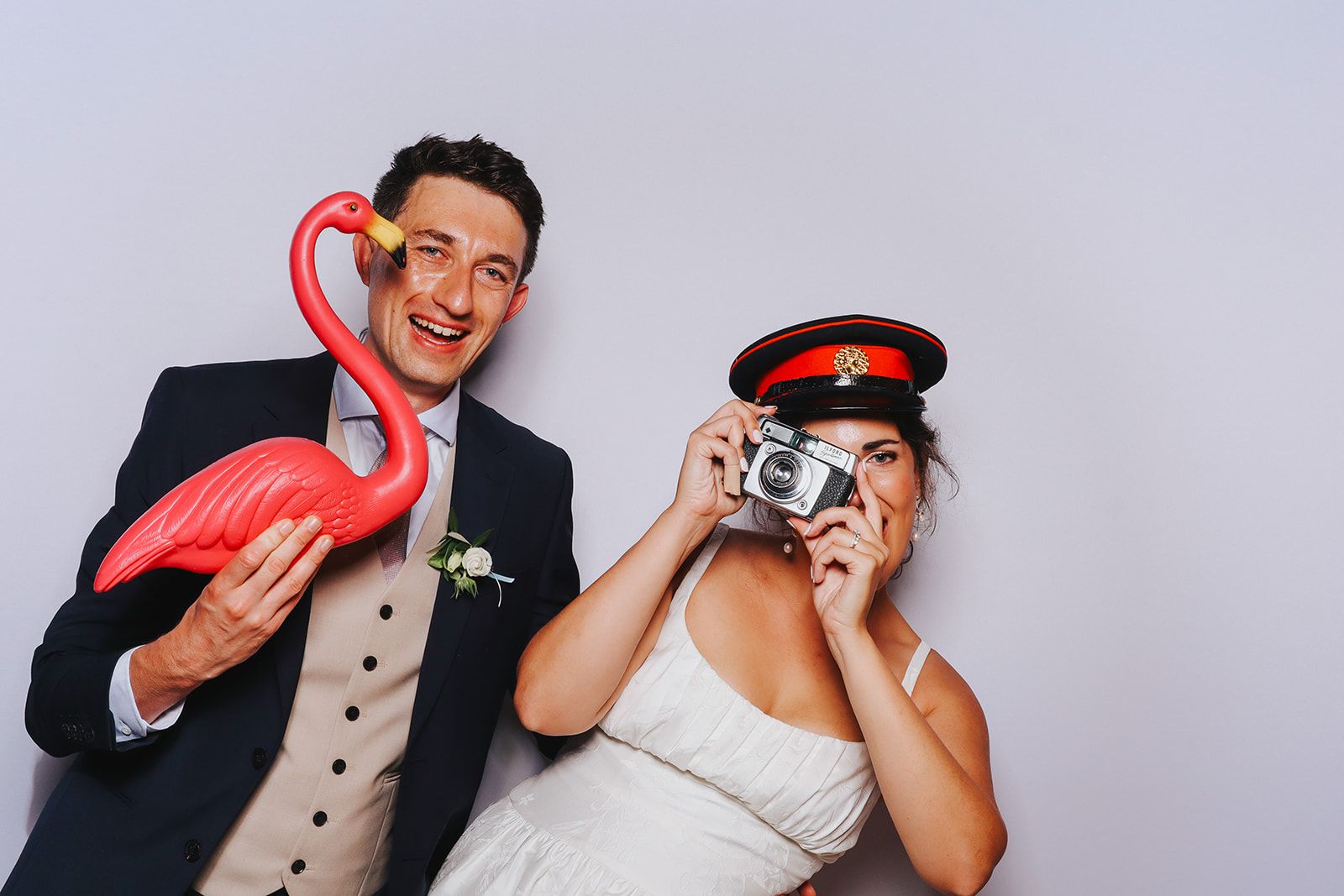 Adrianna and James in the photobooth supplied by The Flash Fox. James is holding a plastic flamingo and Adrianna is taking a photo with an old camera wearing a sailors hat - both are smiling. 