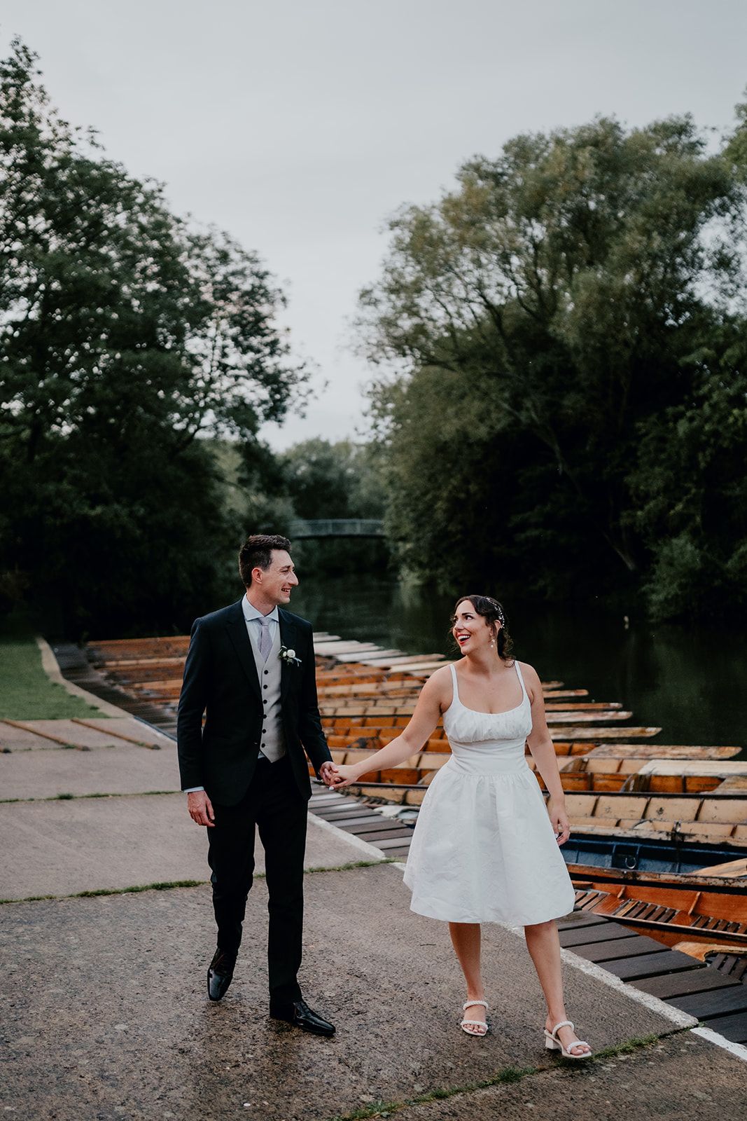 James and Adrianna looking at each other holding hands in the evening light with the punting boats and river in the background. Photo by Matt Fox Photography. 