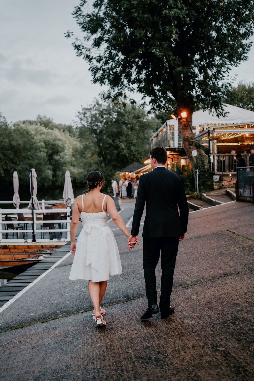 James and Adrianna holding hands looking towards the marquee at Cherwell Boathouse with the lights shining in the evening. Photo thanks to Matt Fox Photography.