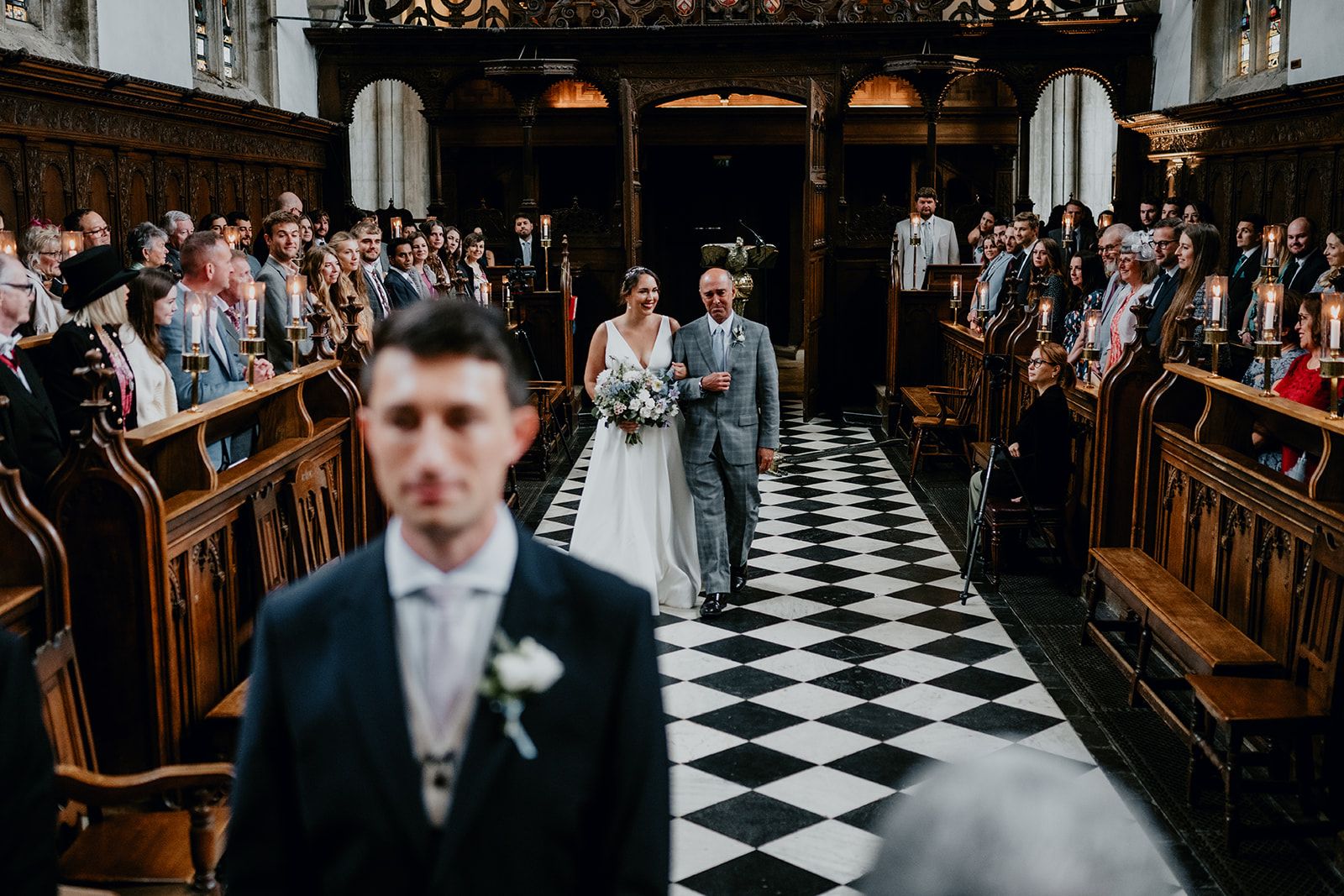 James is out of focus in the foreground, waiting at the top of the alter as Adrianna walks with her Dad down the aisle. Their guests are watching. Photo thanks to Matt Fox Photography.