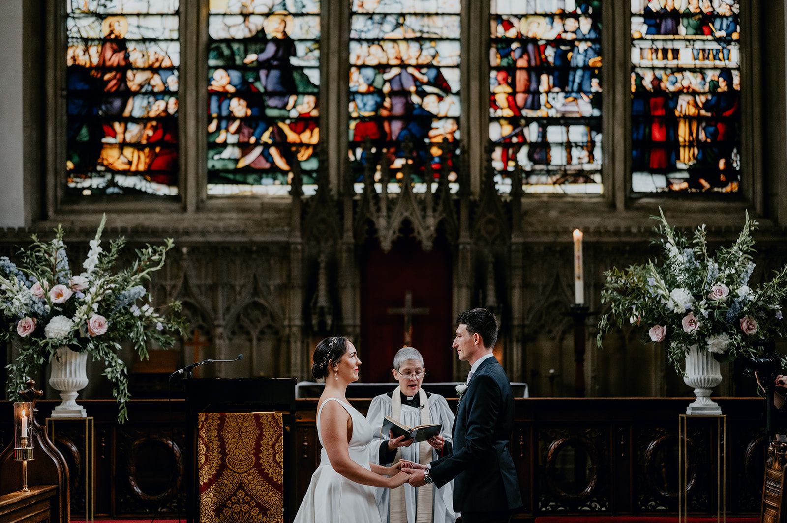 A wide angle photo of James and Adrianna holding hands with the Chaplin leading the ceremony. Flowers frame the shot with the stained glass in the background behind the alter. Photo thanks to Matt Fox Photography.