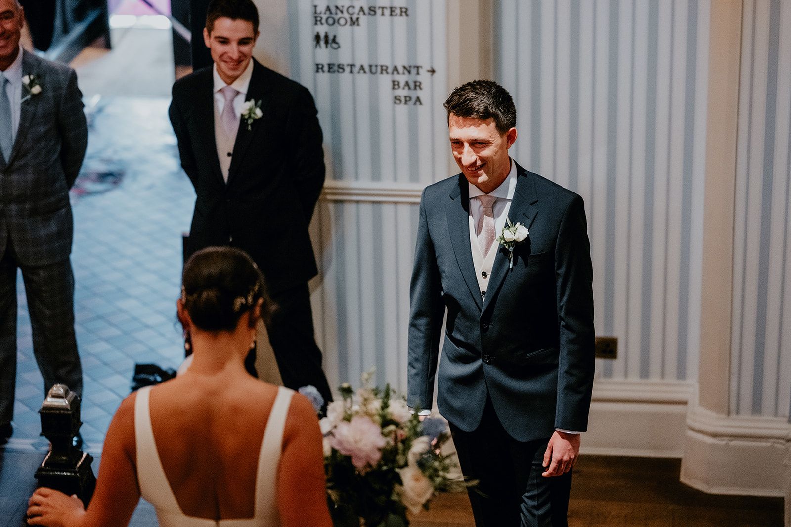 James seeing Adrianna in her wedding dress for the first time. He's smiling as she approaches him. His best man is also smiling in the background. Photo thanks to Matt Fox Photography.