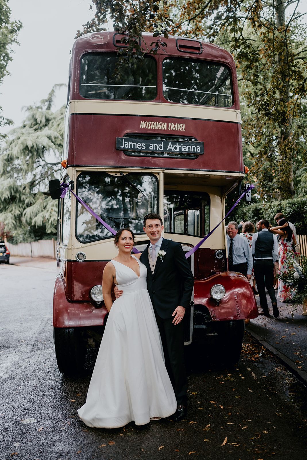 Adrianna and James stood in front of the vintage red bus outside Cherwell Boathouse entrance as their guests walk to the reception in the background. Photo courtesy of Matt Fox Photography.