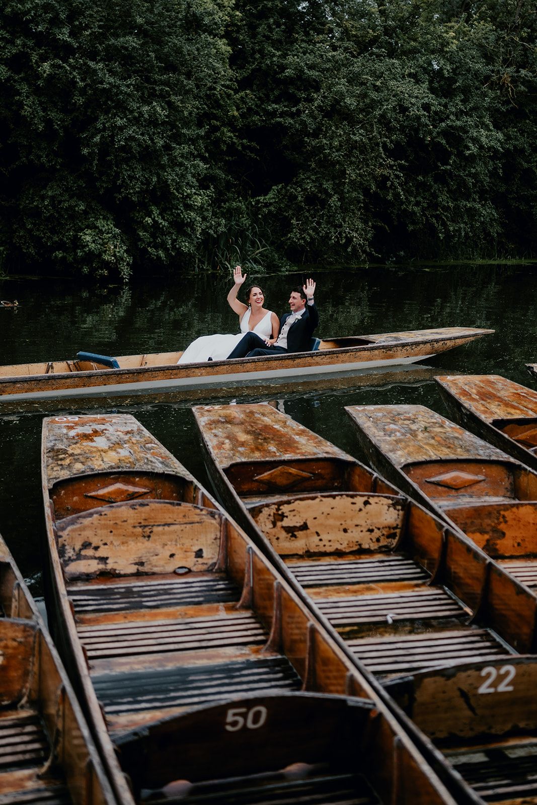 James and Adrianna wave to their guests onboard a punting boat. Empty punts are in the foreground on the river. Photo courtesy of Matt Fox Photography