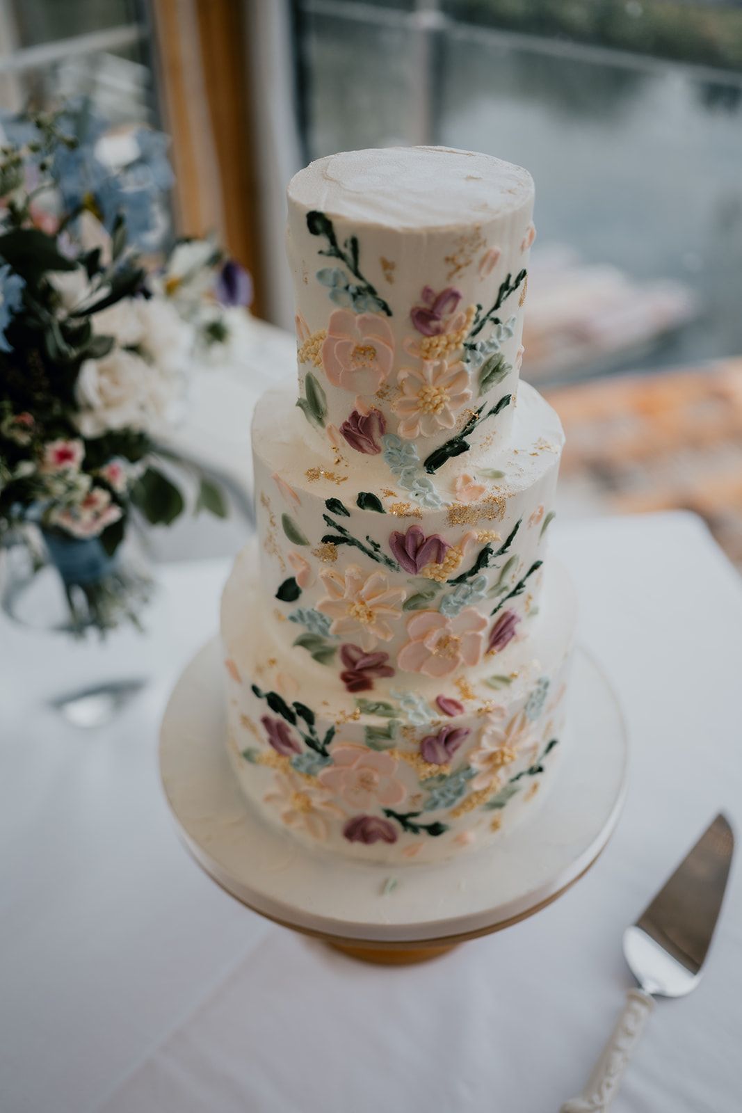 Beautiful three tier wedding cake with flowers made out of icing all down the front. A cake knife is in the foreground and flowers in the background are out of focus. Photo courtesy of Matt Fox Photography.