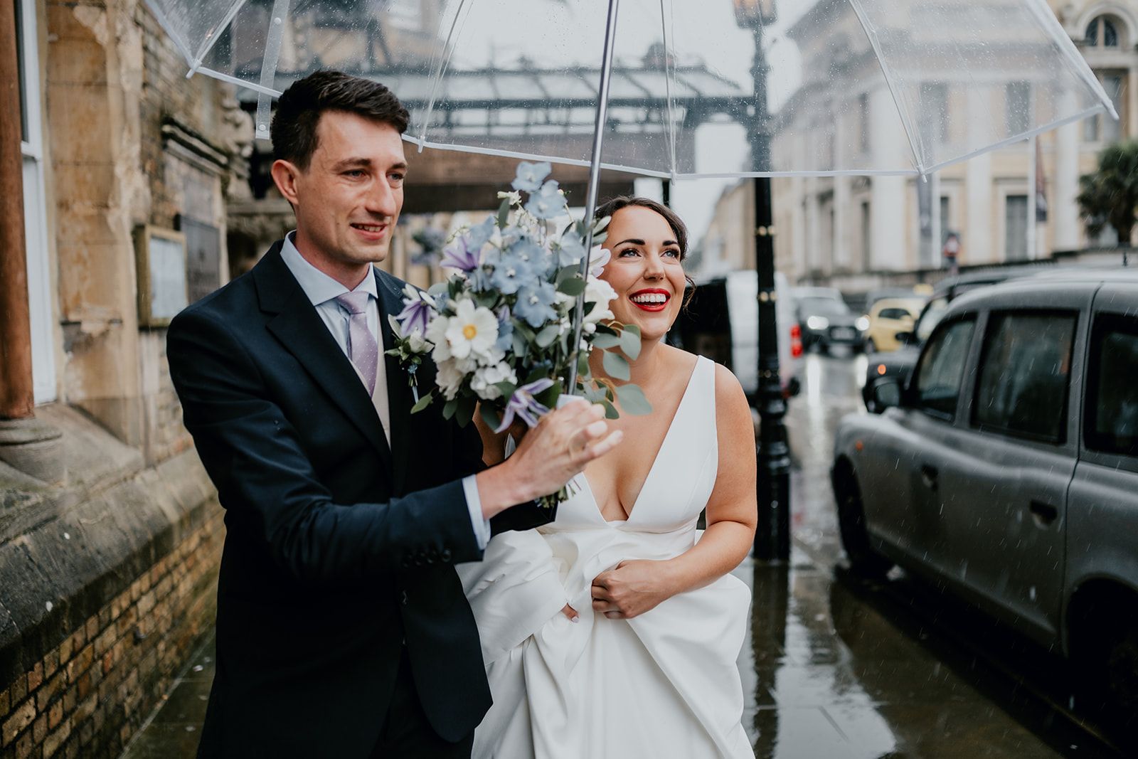 James holding an umbrella in front of The Randolph Hotel in Oxford whilst Adrianna holds up her wedding dress - she is looking up at the rain smiling. A taxi and other cars are in the background. Photo thanks to Matt Fox Photography.